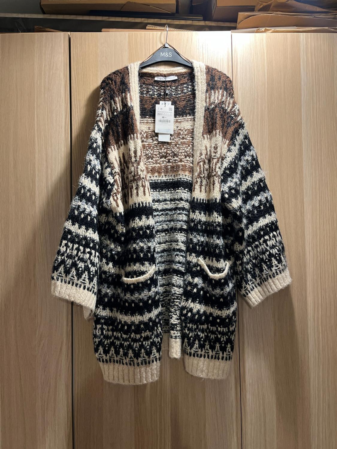 Chunky knit cardigan with a geometric pattern in black, white and brown, hanging in a wooden wardrobe. Texts: M&S, zara.