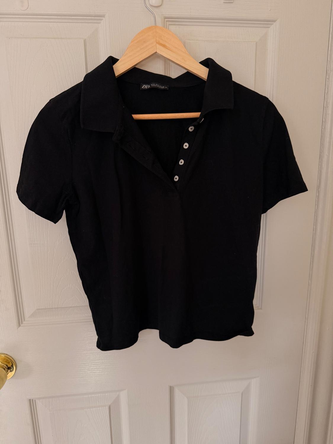 Black short-sleeved polo shirt hanging on a wooden hanger over a white door. Silver buttons visible on the front.