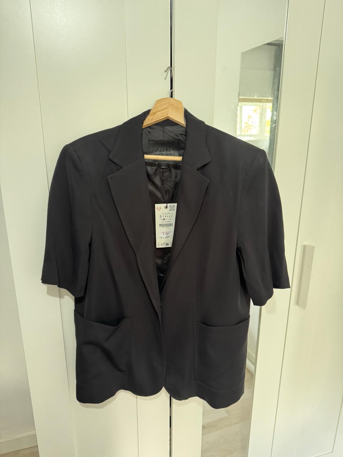 Μαύρο blazer με κοντά μανίκια και τσέπες, κρεμασμένο σε ξύλινη κρεμάστρα μπροστά από μια λευκή ντουλάπα. Ορατή εσωτερική ετικέτα zara.