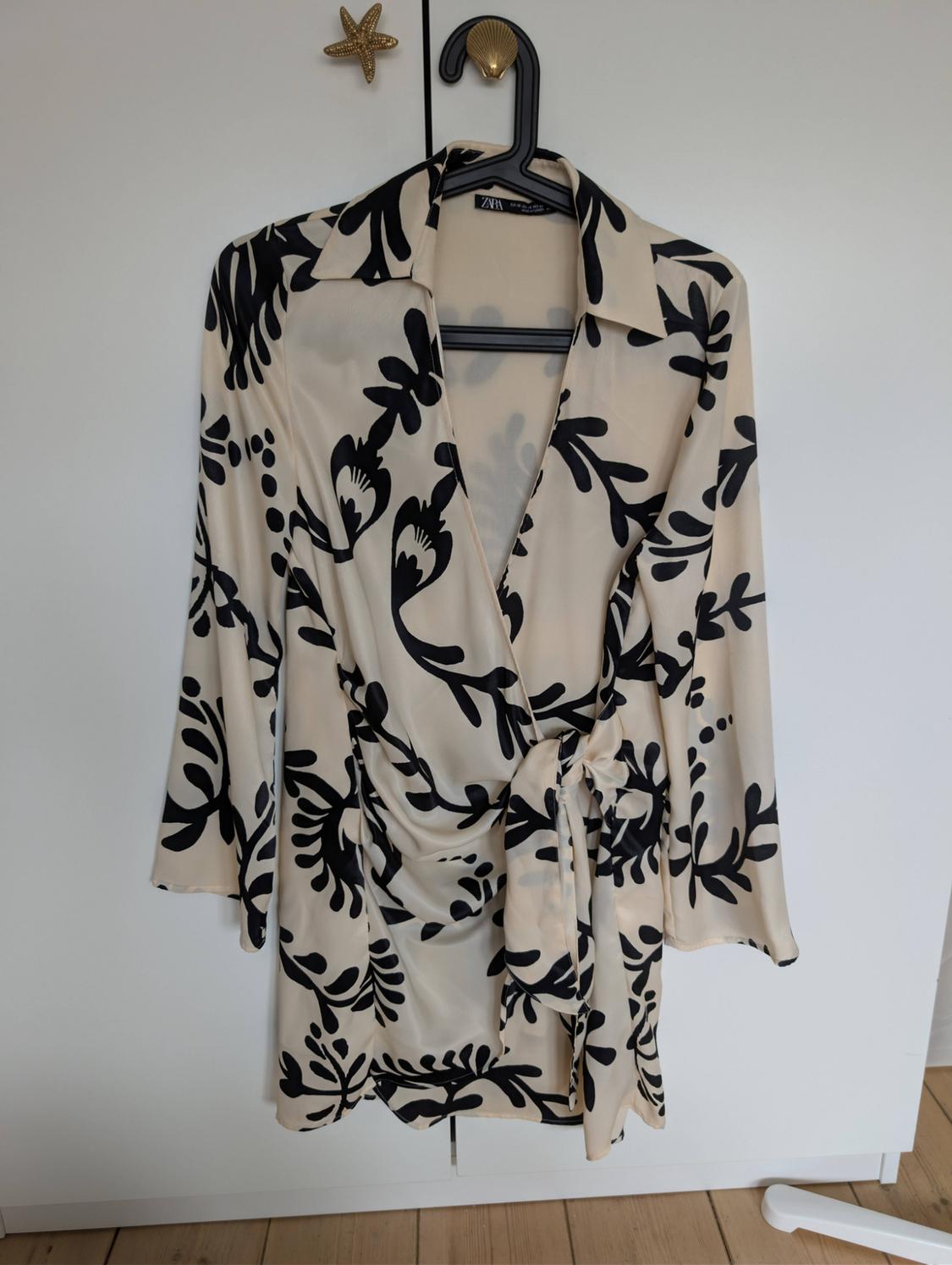 Beige wrap dress with a black leaf print and tie waist, hanging in a white wardrobe. Text on the label: ZARA.