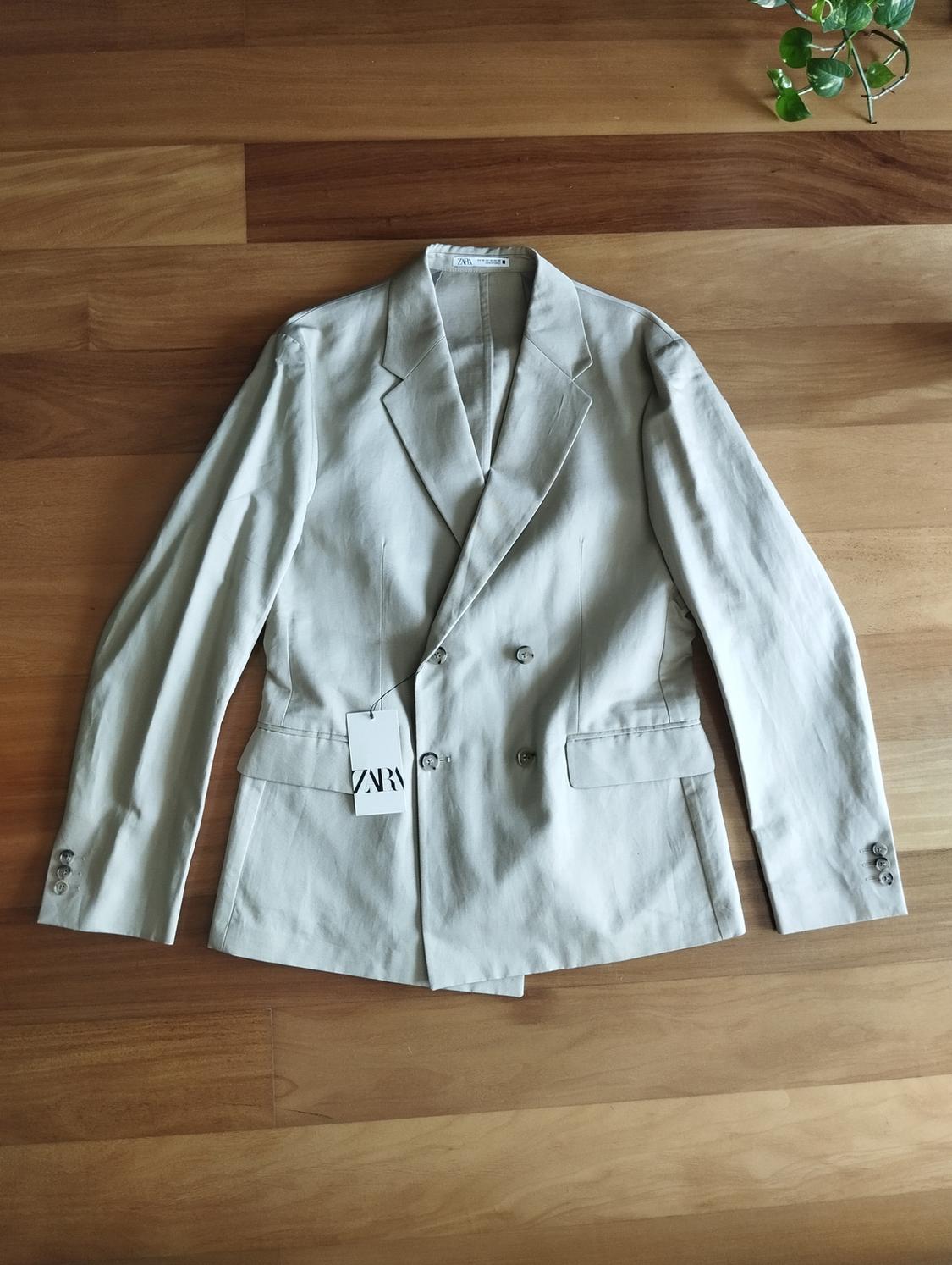 Chaqueta beige de doble botonadura sobre suelo de madera, con etiqueta colgante que dice "ZARA". Planta verde en la esquina superior derecha.
