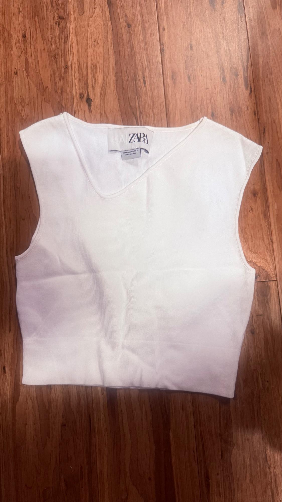 White sleeveless cropped top with an asymmetric neckline on a wooden background. Label with the text KAIA ZARA.