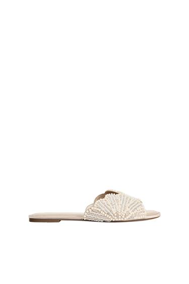 Elegant beige flat sandal seen in profile against a white background. Its wide strap is decorated with pearls and shiny beads.