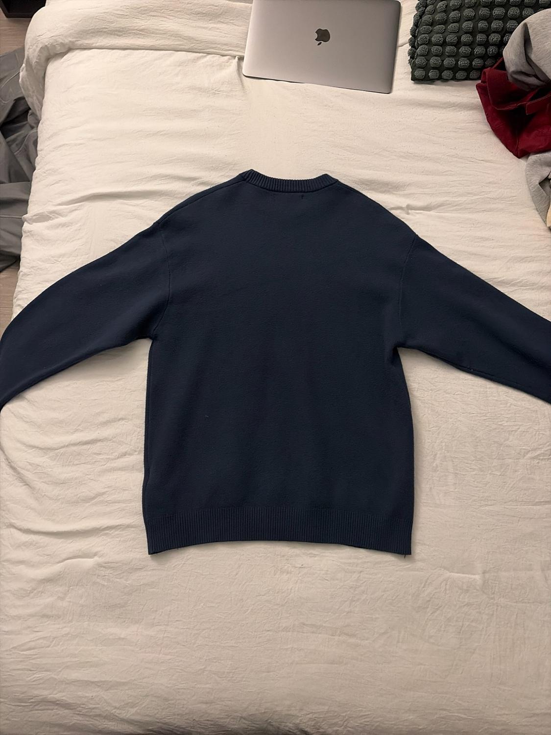Navy blue sweater spread out on a white bed with a closed silver laptop, conveying a relaxed and casual vibe.