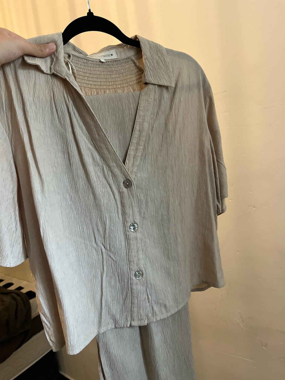 Hand holding a hanger with a beige crinkled-texture outfit, consisting of a short-sleeved button-up shirt and trousers.