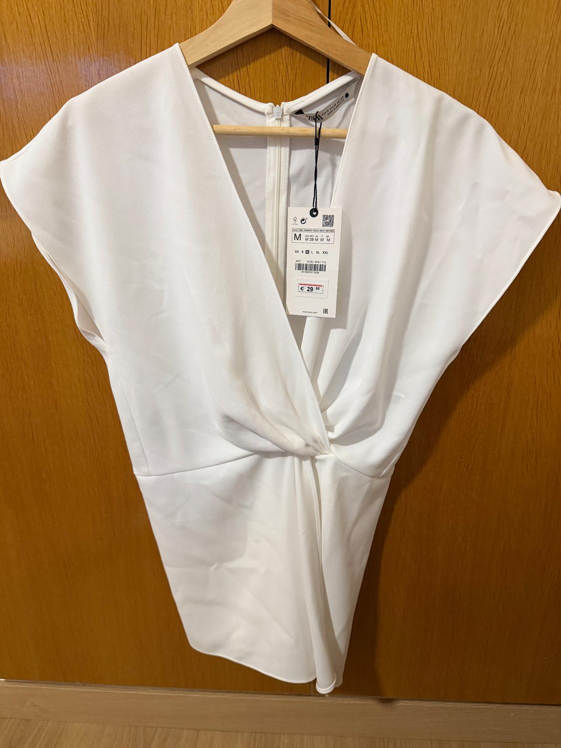 White dress with V-neckline and pleats, hanging on a wooden hanger. Visible tag with price of €29.99. Wooden background.