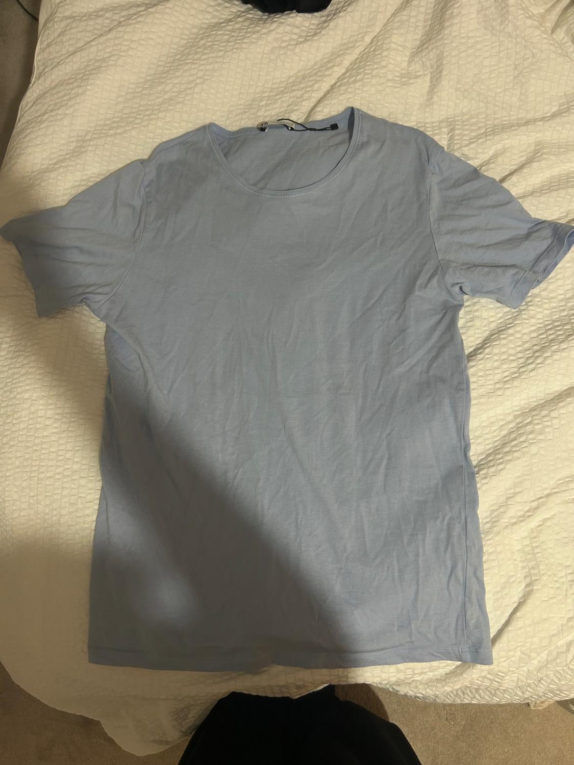Wrinkled light blue t-shirt on a bed with a textured white bedspread. Part of a dark garment visible at the top.
