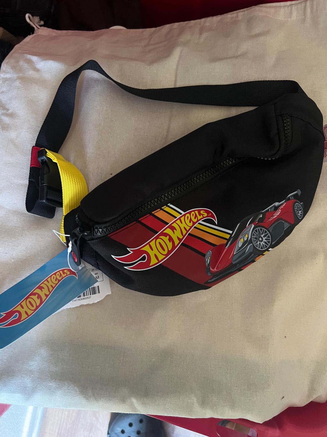 Black bum bag on light-coloured fabric. Printed with the text Hot Wheels, a red car and stripes. Black strap with yellow detail and blue label.