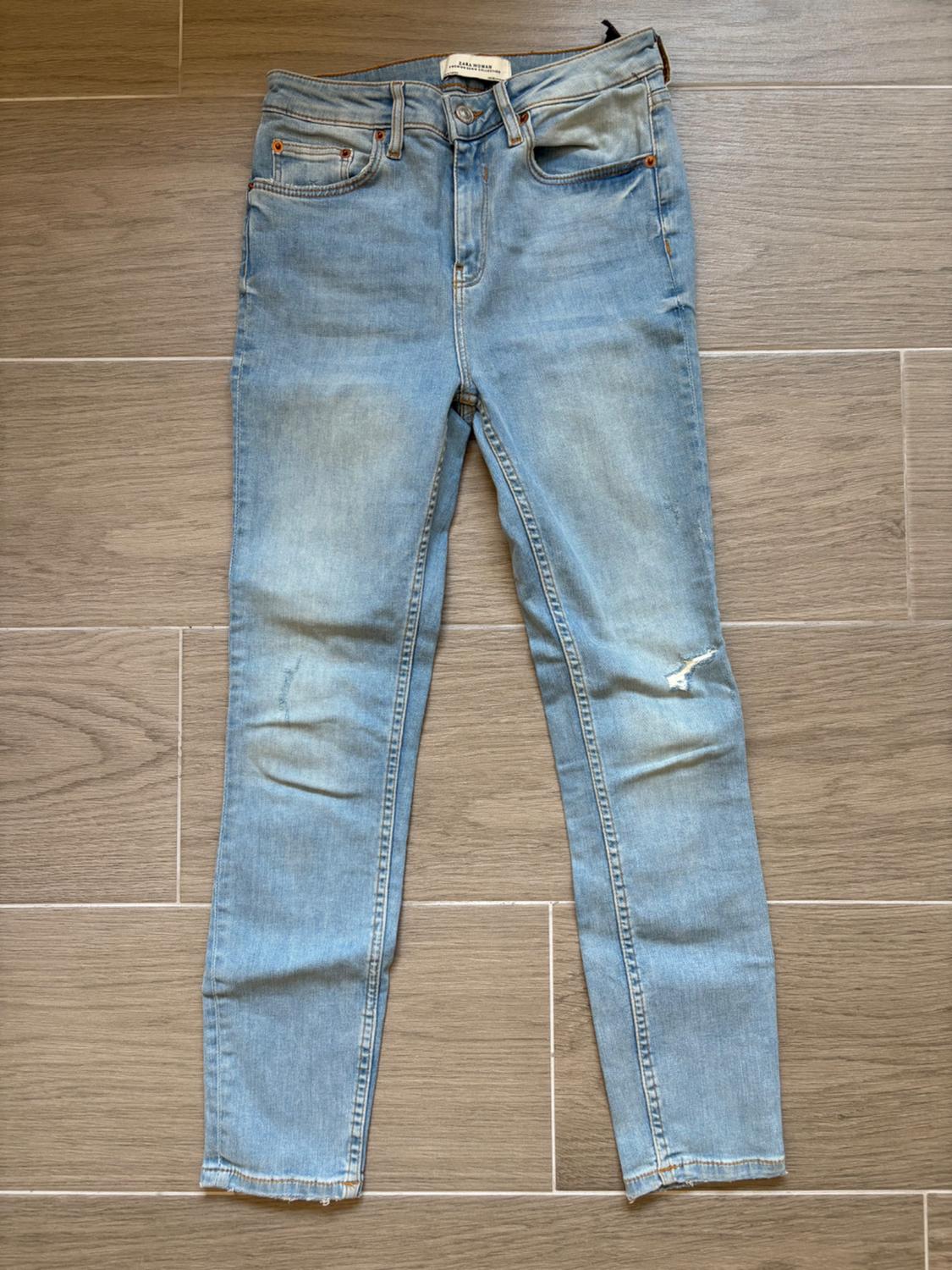 Light blue jeans with a distressed effect, rip on the right leg, raw hem, on light brown wooden floor.
