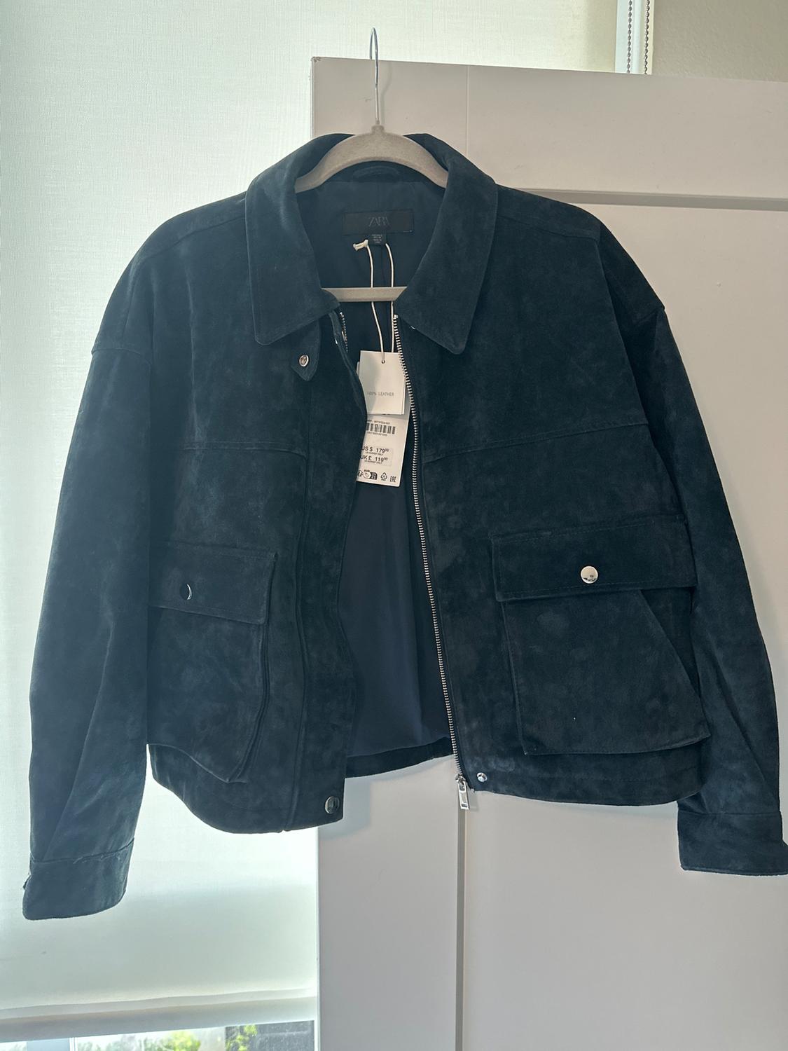 Black suede jacket hanging on a hanger, with front pockets and a zipper, against a light background. Visible tags.