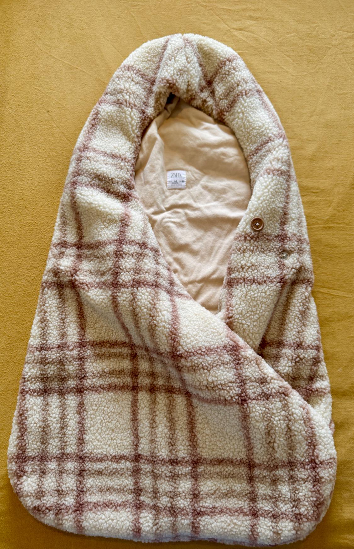 Cream and brown checked soft fabric baby sleeping bag with hood. Plain interior with ZARA label. Button fastening, on a yellow background.