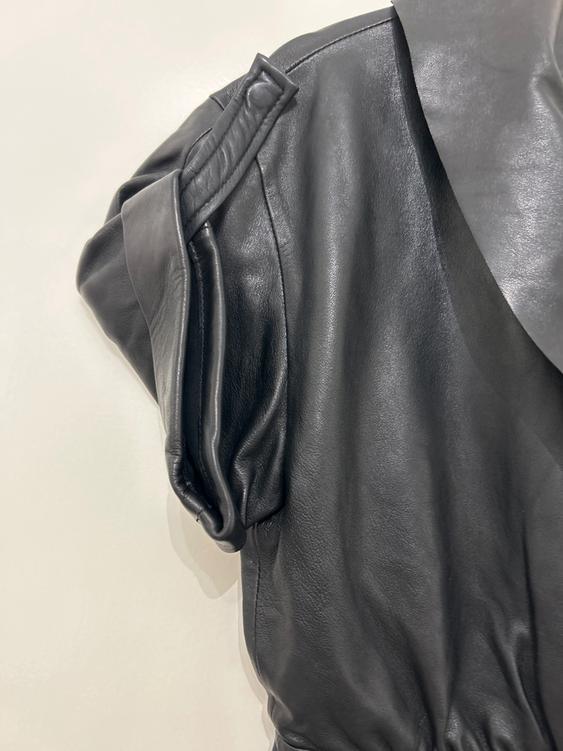 Detail of a black leather jacket with shoulder pad and pleats, on a plain white background.