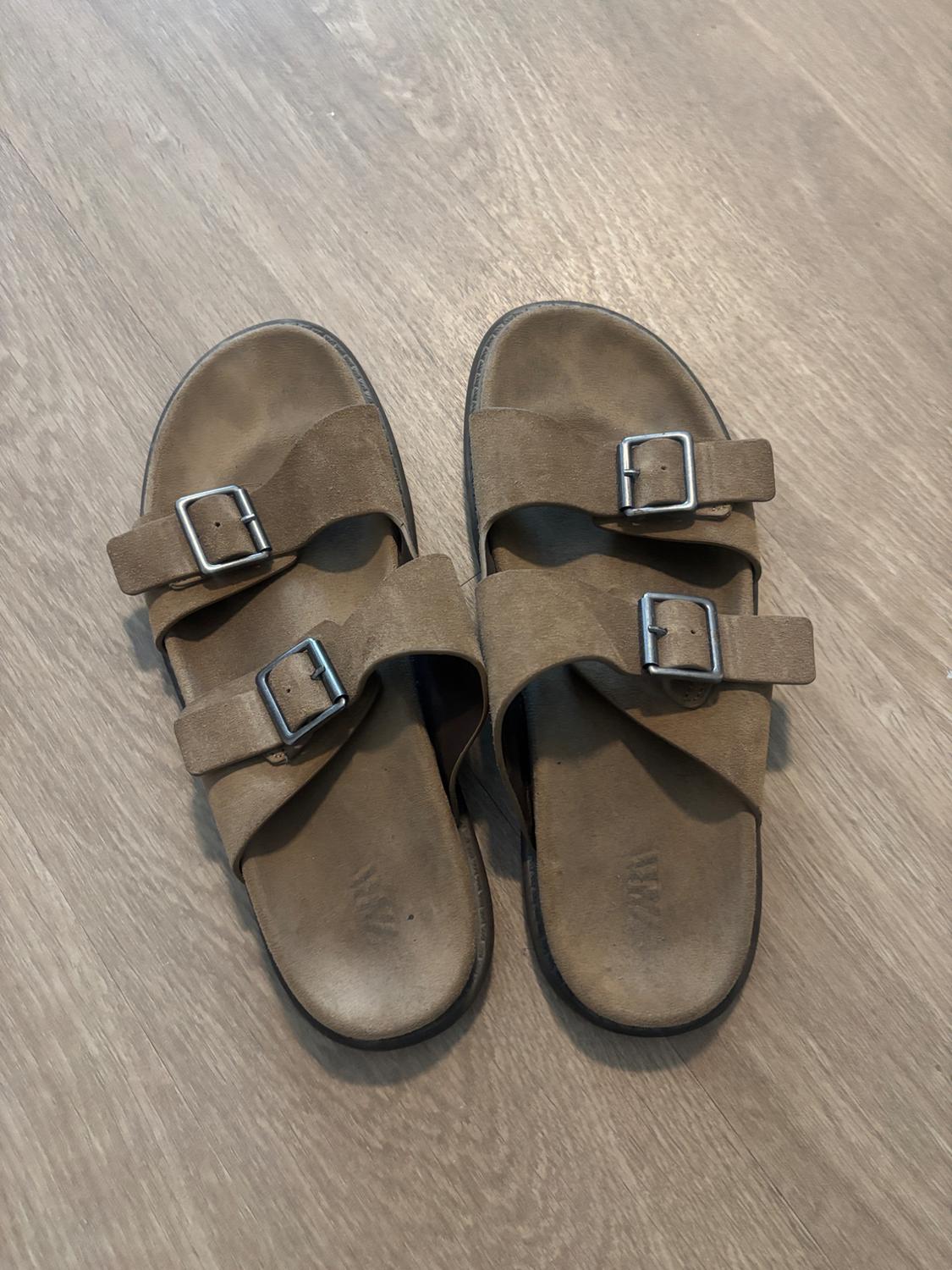 Pair of brown double-strap sandals with buckles on a light wood floor. ZARA text on the insole.