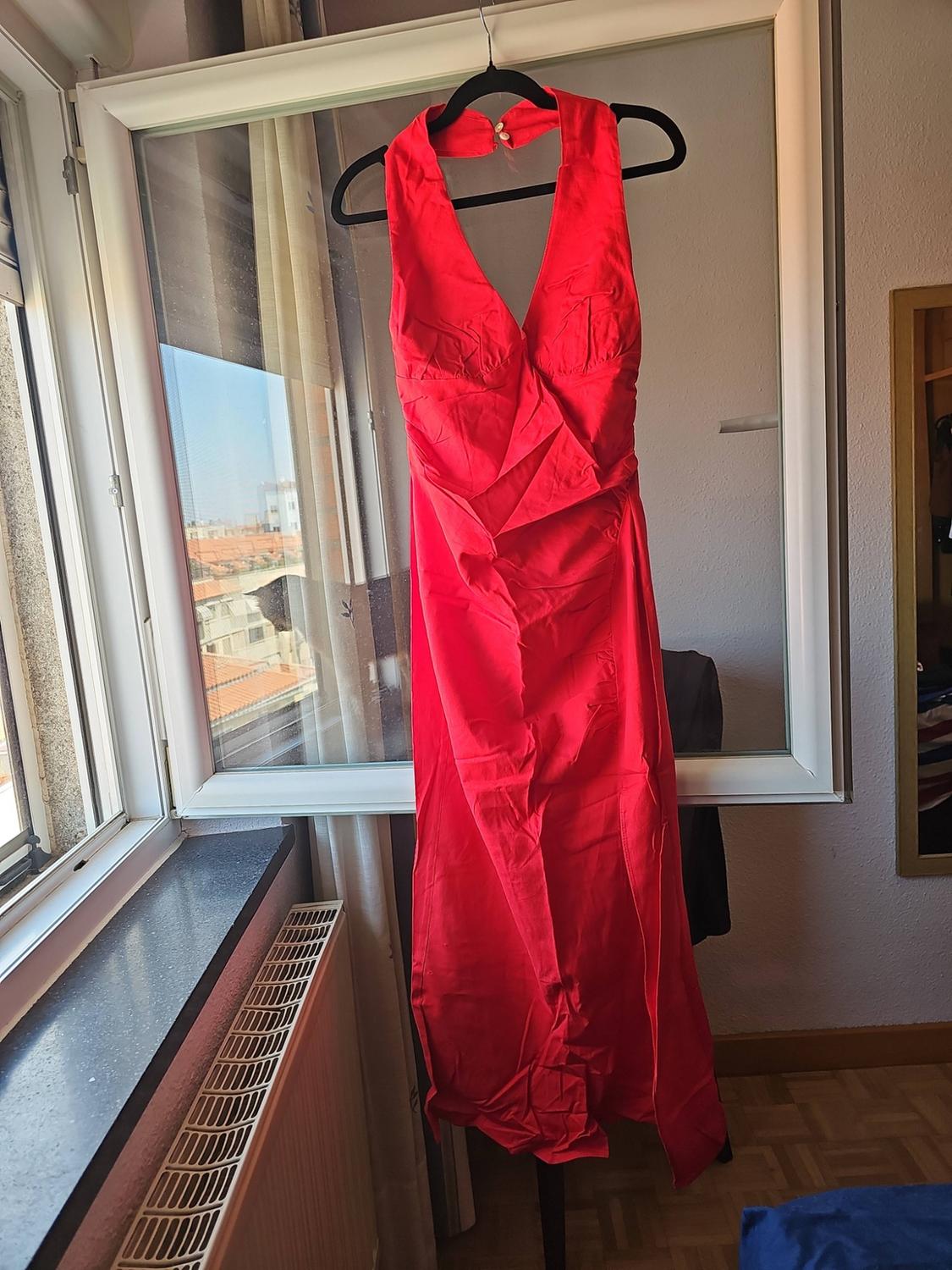 Κόκκινο μακρύ φόρεμα με δέσιμο στο λαιμό (halter neck), λαιμόκοψη V, σούρες και άνοιγμα, κρεμασμένο σε κρεμάστρα μπροστά από ένα ανοιχτό παράθυρο.