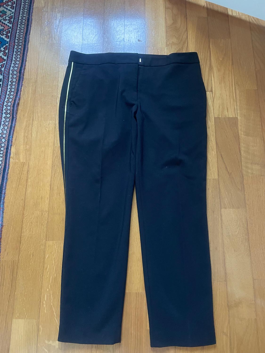 Black trousers with a yellow side stripe on a wooden floor, next to a rug with a geometric pattern on the left edge.