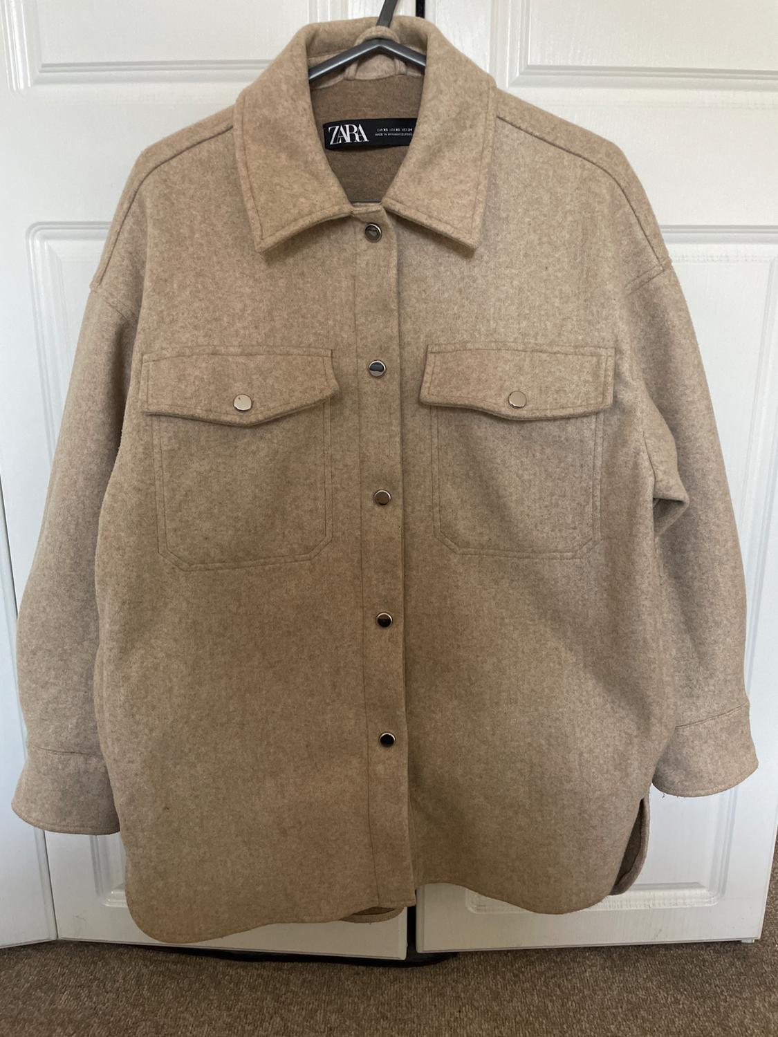 Beige overshirt with buttons and chest pockets, hanging on a hanger in front of a white door. ZARA text on the label.