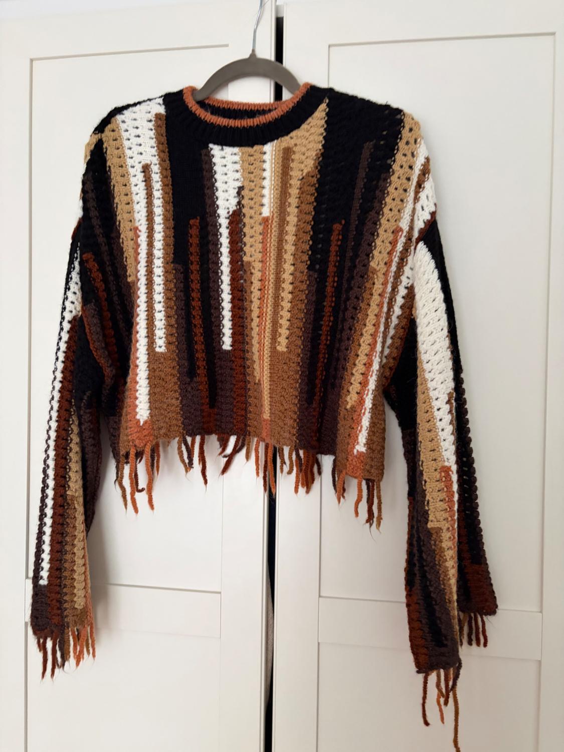 Multicoloured knitted jumper with fringes hanging on a hanger in front of a white wardrobe. Brown, black, and beige tones.