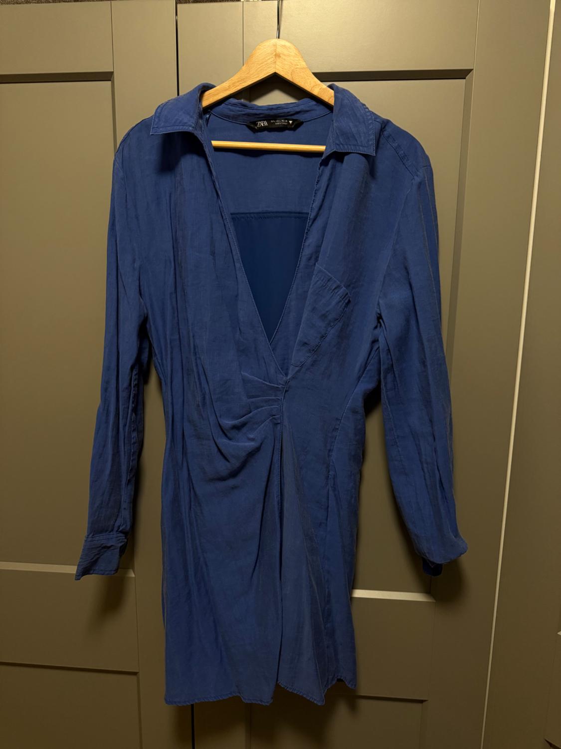 Long-sleeved blue dress with a V-neckline and draping, hanging on a wooden hanger in front of a wardrobe. Tag with the text ZARA.