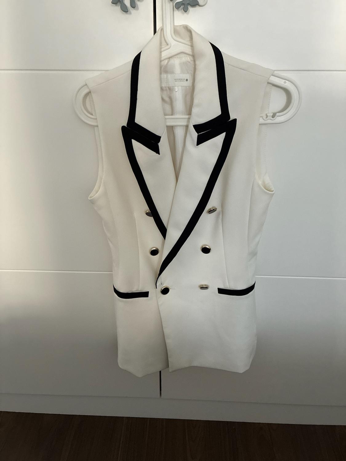 White sleeveless waistcoat with black trim lapels and double-breasted buttons, hanging on a hanger in front of a white wardrobe.