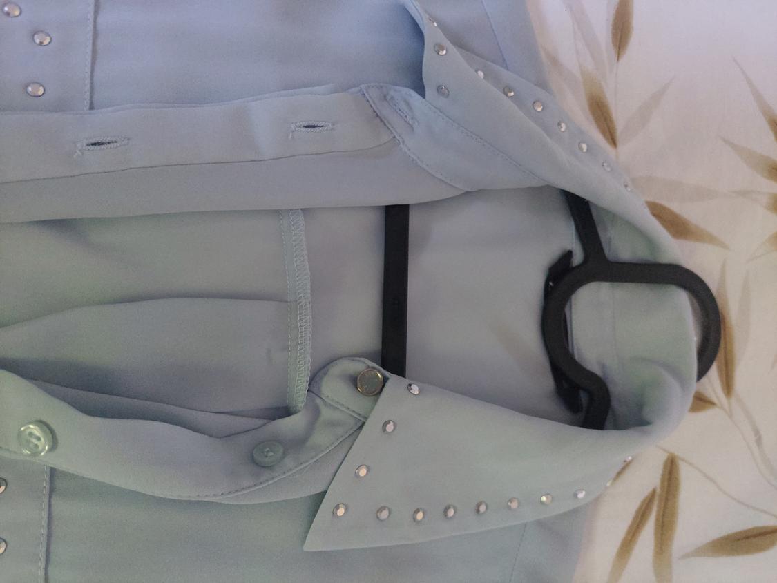 Light blue shirt with buttons and stud details on the collar, hanging on a black hanger on a surface with leaves.