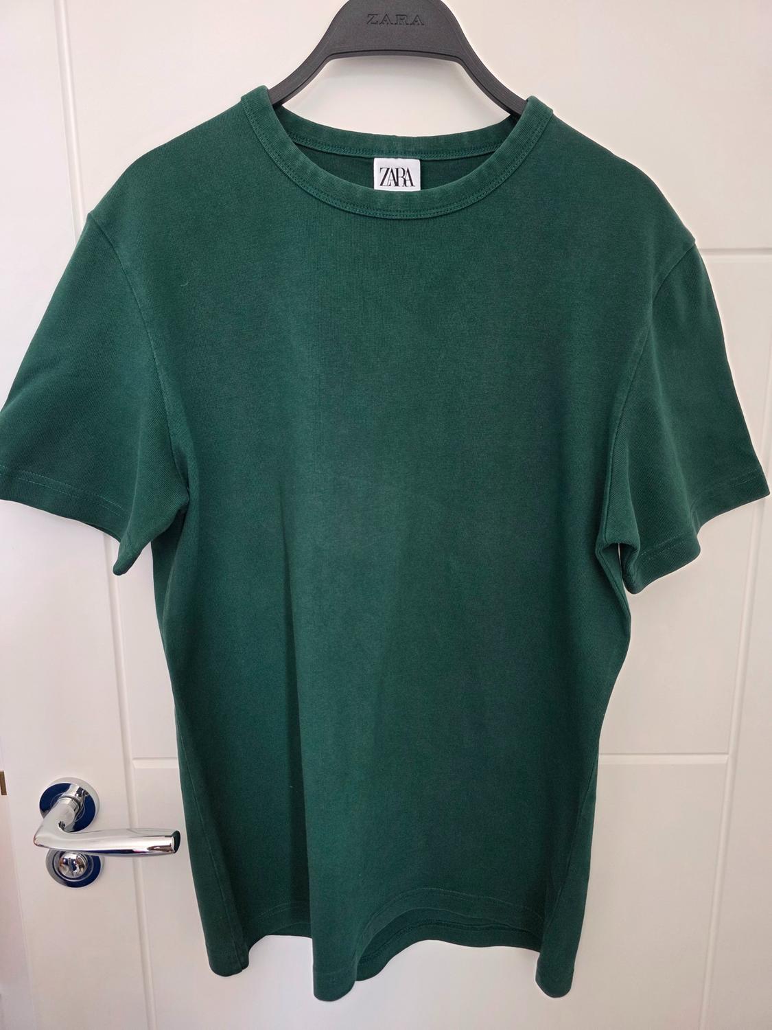 Dark green short-sleeved T-shirt on a black hanger on a white door with a silver handle. ZARA text visible on the label and the hanger.