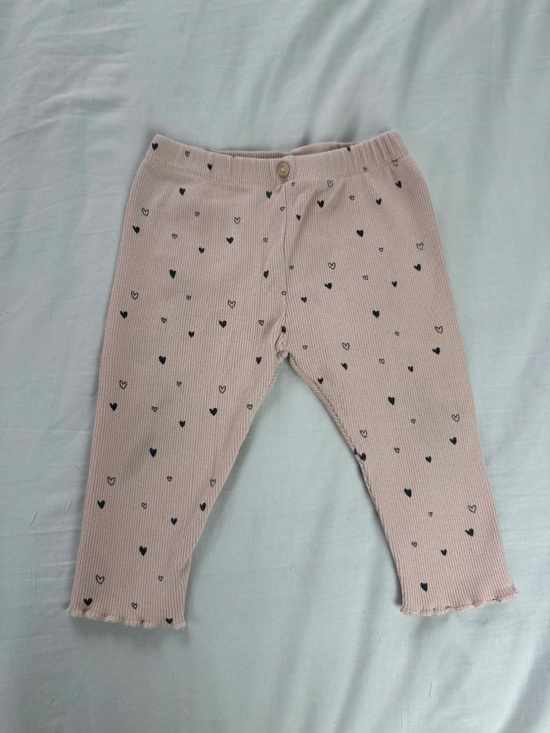 Light pink ribbed leggings with an all-over print of small black hearts, with a button at the waist, on a white fabric background.