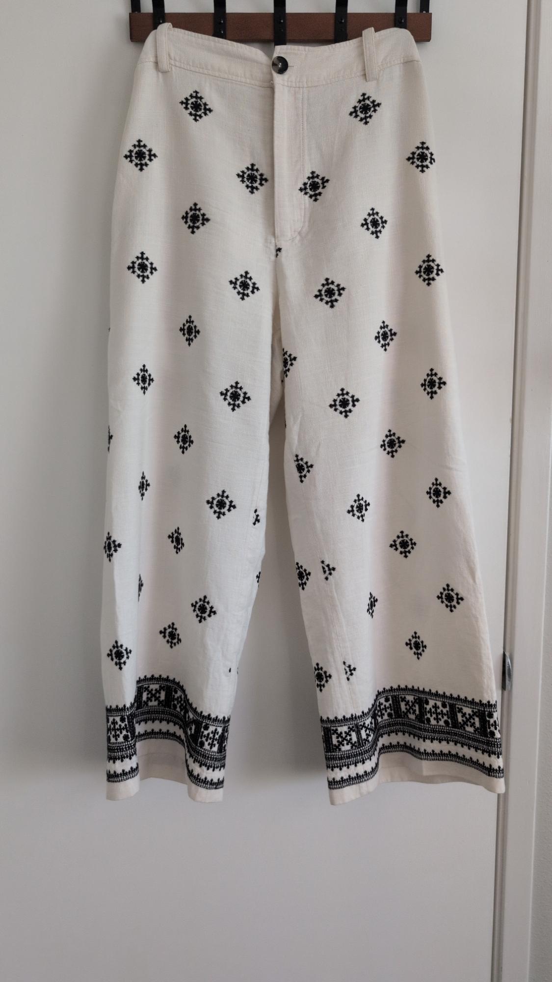 Wide white trousers with black embroidery and a decorative border on the hem, hanging on a hanger over a white door.