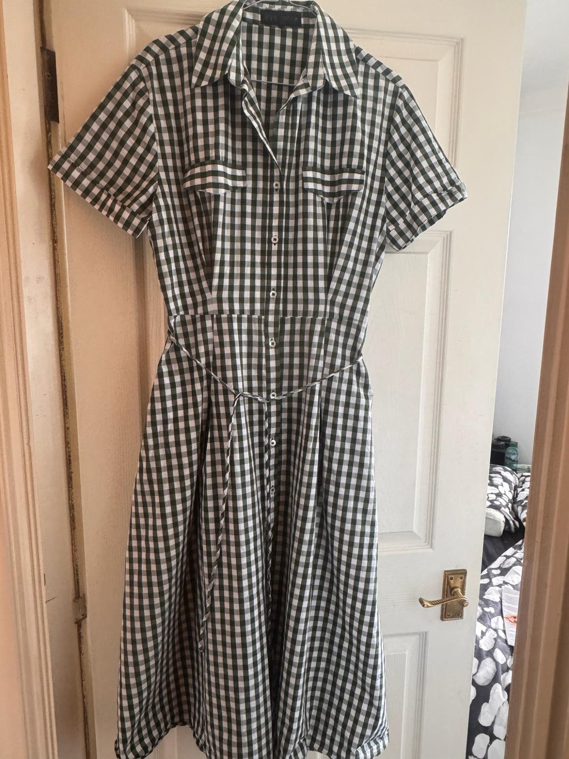 Green-and-white checked shirt dress with buttons, hanging on a partly open white door that reveals a bedroom.