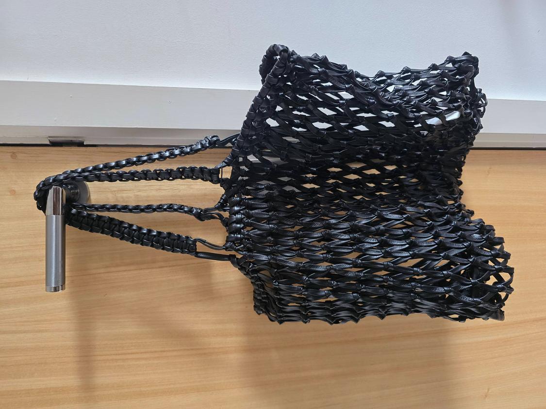 Black woven handbag hanging from a metal hook on a white wall and light wood background.