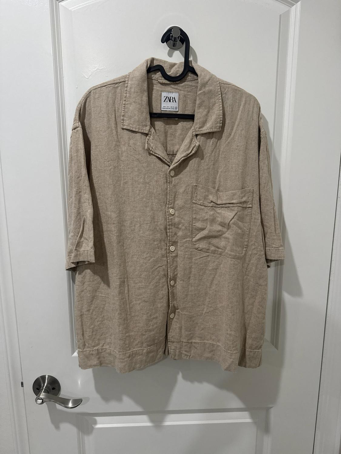 Beige short-sleeved shirt with buttons and a pocket, hanging on a hanger on a white door. Text: ZARA.
