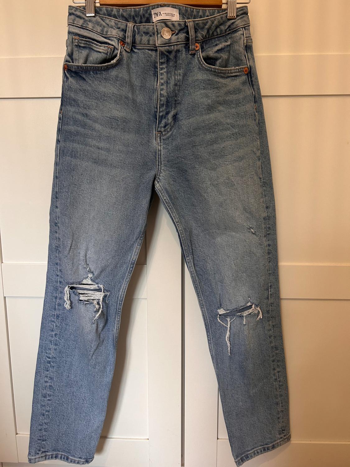 Worn blue jeans with ripped knees, hanging on a hanger against a background of white panels.