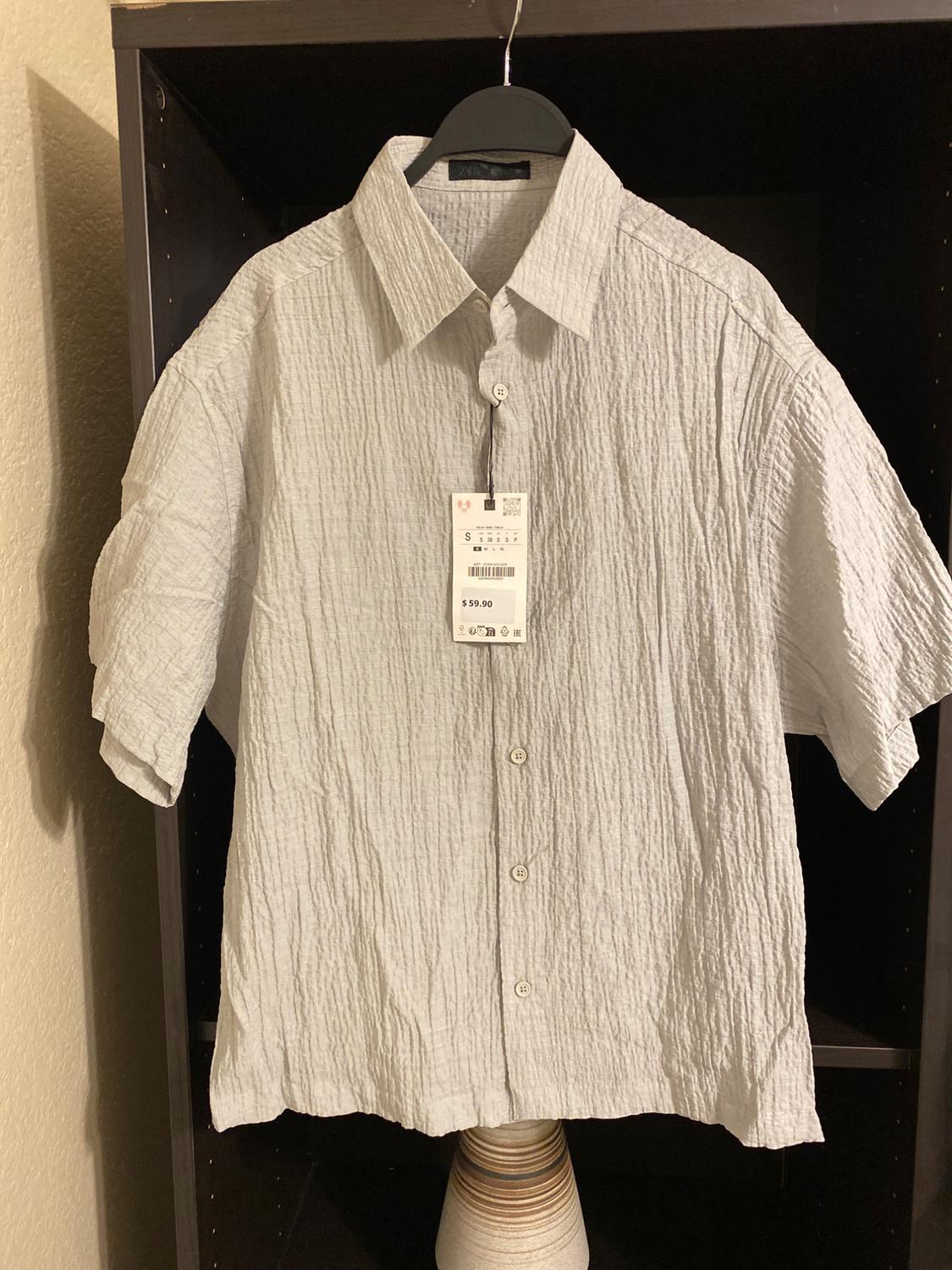 White short-sleeved shirt with a crinkled texture, hanging on a black hanger. Visible tag with a price of $59.90. Dark background.