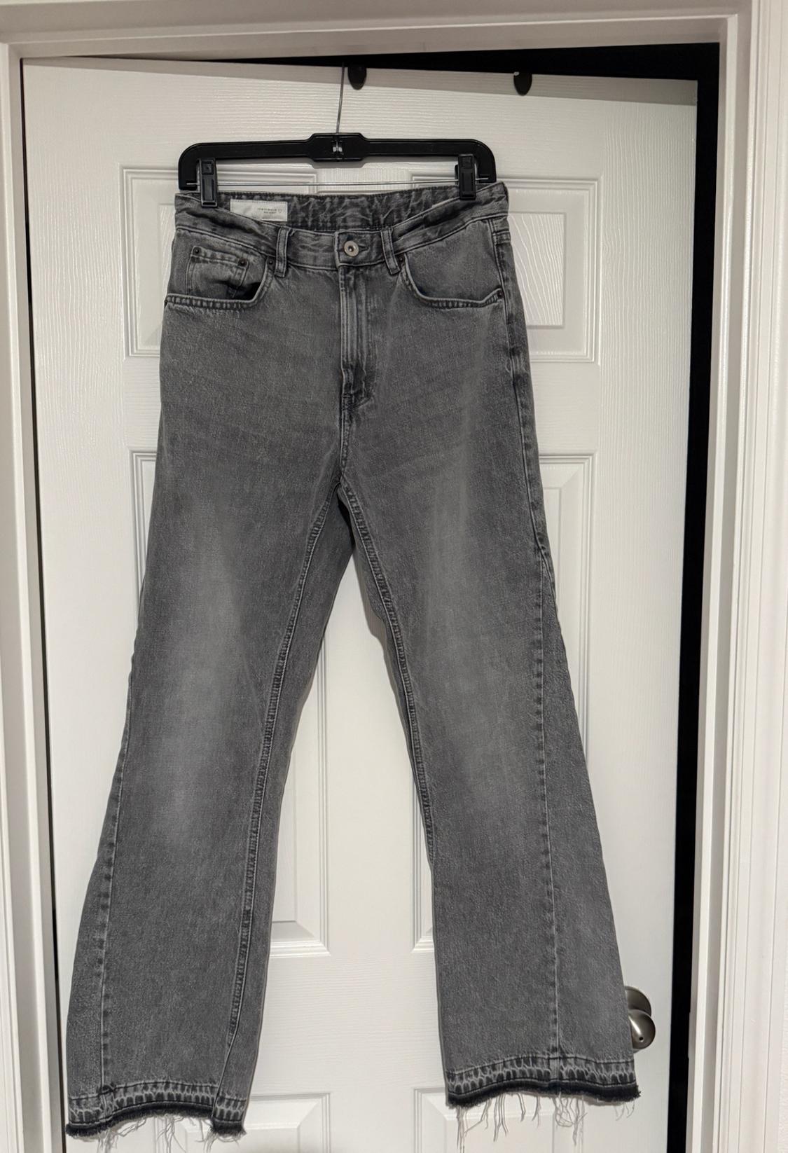 Gray jeans hanging from a hanger on a white door. They have a distressed wash and a frayed hem with a darker stripe.