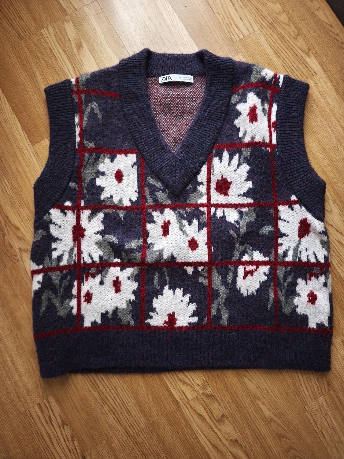 Sleeveless knitted waistcoat with a checked design and white and red flowers on a blue background, on a light wooden floor.