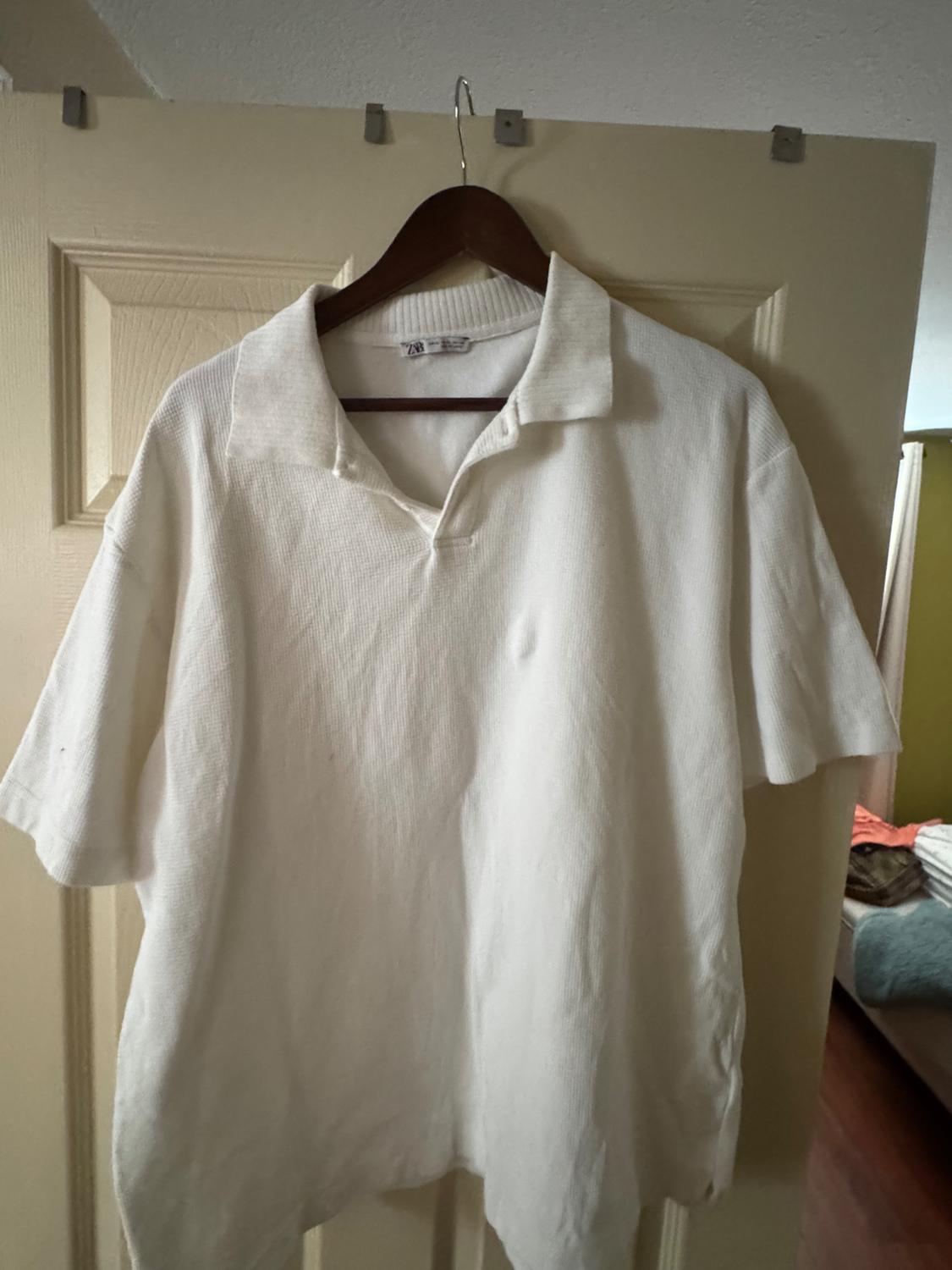 Textured white short-sleeve polo shirt hanging on a wooden hanger on a door. ZARA label. Casual home setting.