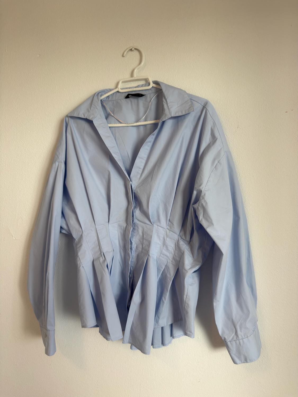 Light blue long-sleeved button-up shirt with a pleated waist, hanging on a white hanger against a plain white wall.