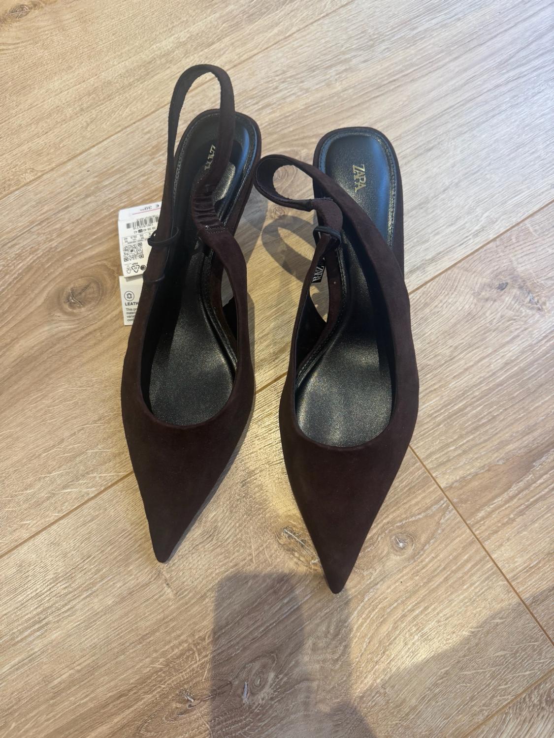Dark brown slingback high-heeled shoes with a pointed toe and label, on a light wood floor. Text: ZARA.