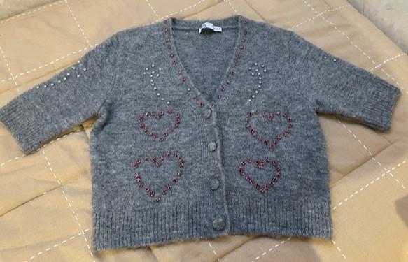 Short-sleeved grey cardigan with buttons and red embroidered hearts. Beige fabric background with white lines.