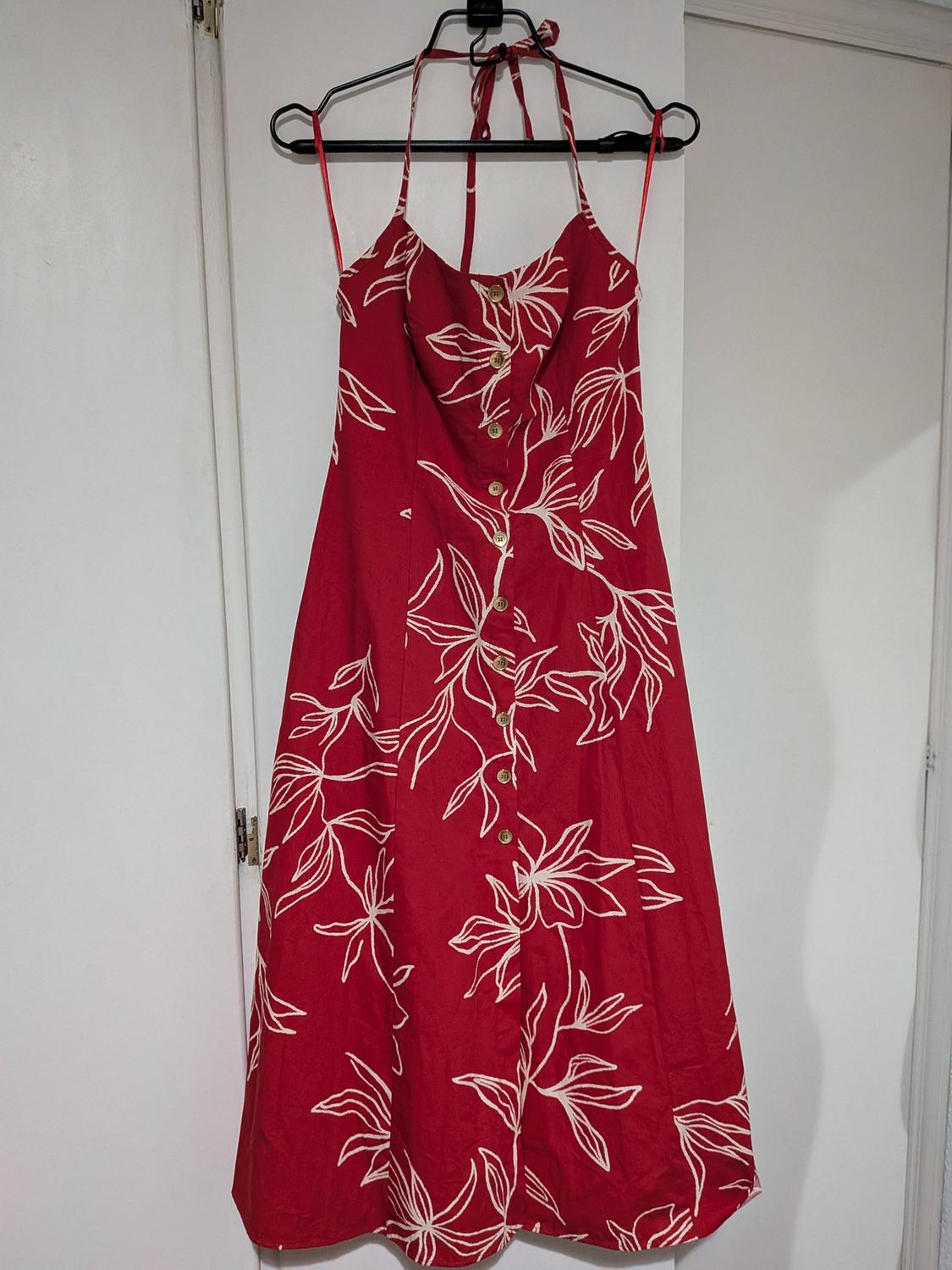 Red dress with white leaf print, halter neck and front buttons, hanging on a black hanger on a white door.