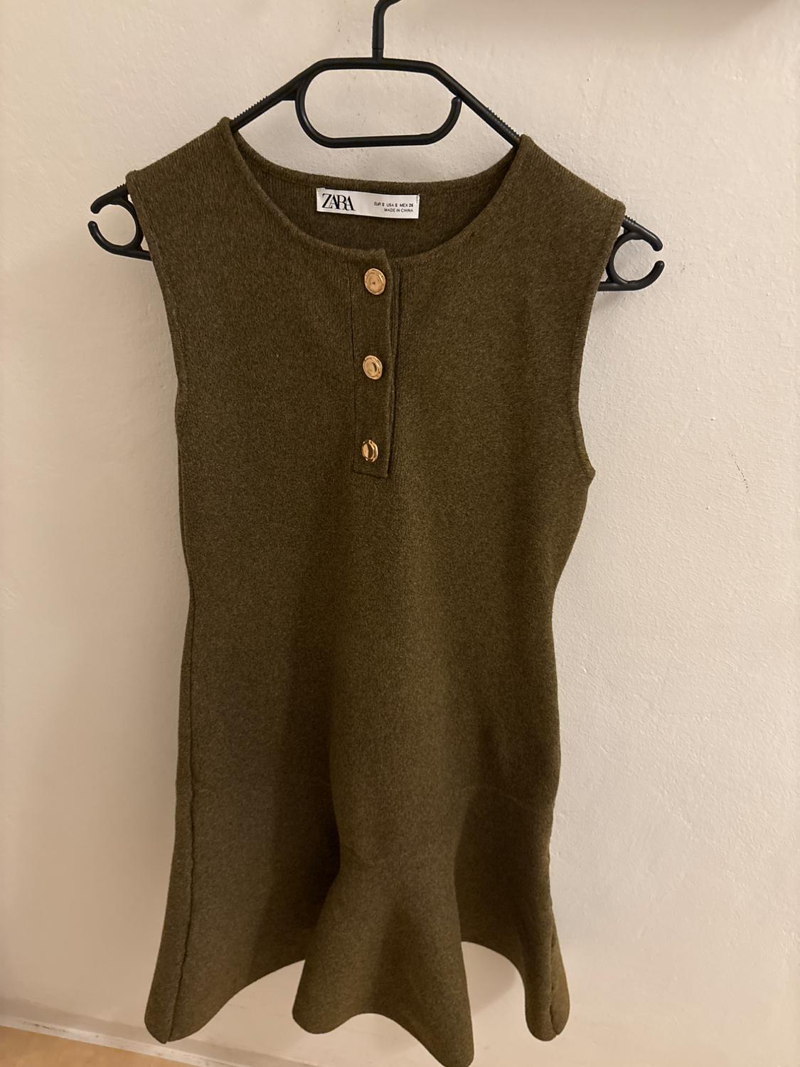 Olive green sleeveless dress with wooden buttons on the chest, hanging on a black hanger against a white wall.