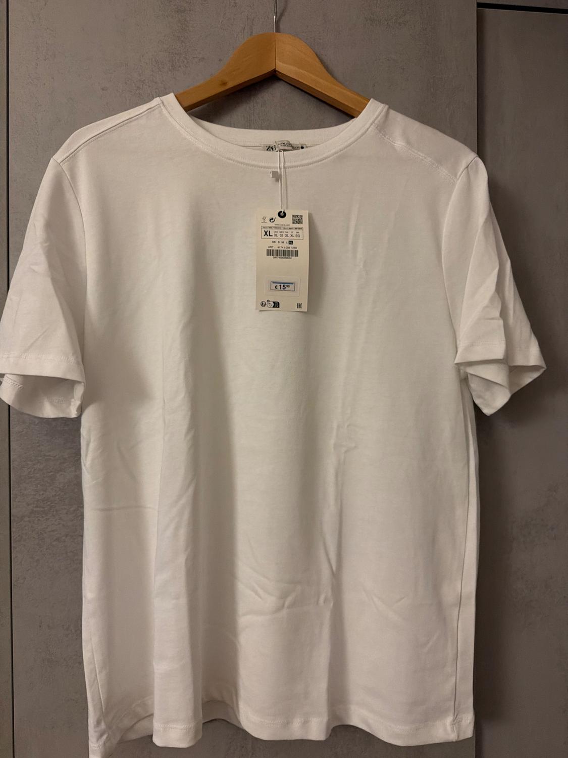 White, short-sleeved T-shirt hanging on a wooden hanger. Visible label with size XL and price of 15 euros. Grey background.
