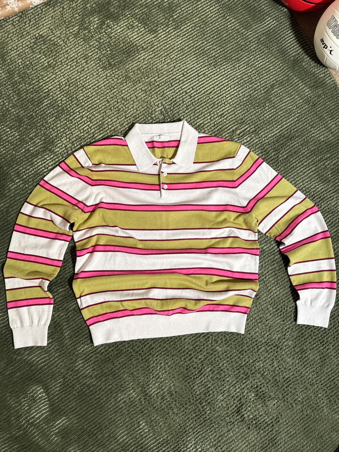 Green, pink, and white horizontal striped jumper on a green rug. Long sleeves and a button-up polo neck.