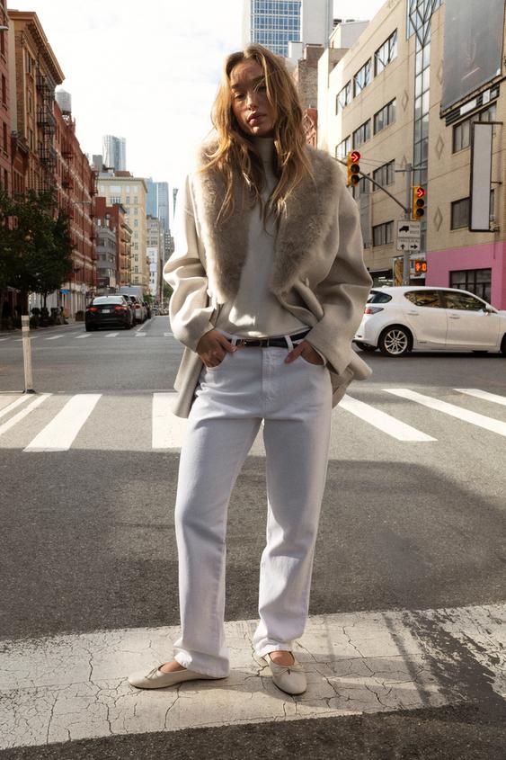 Sada pískového kabátu s límcem s efektem kožešiny, světlým svetrem, bílými kalhotami a béžovými plochými botami.