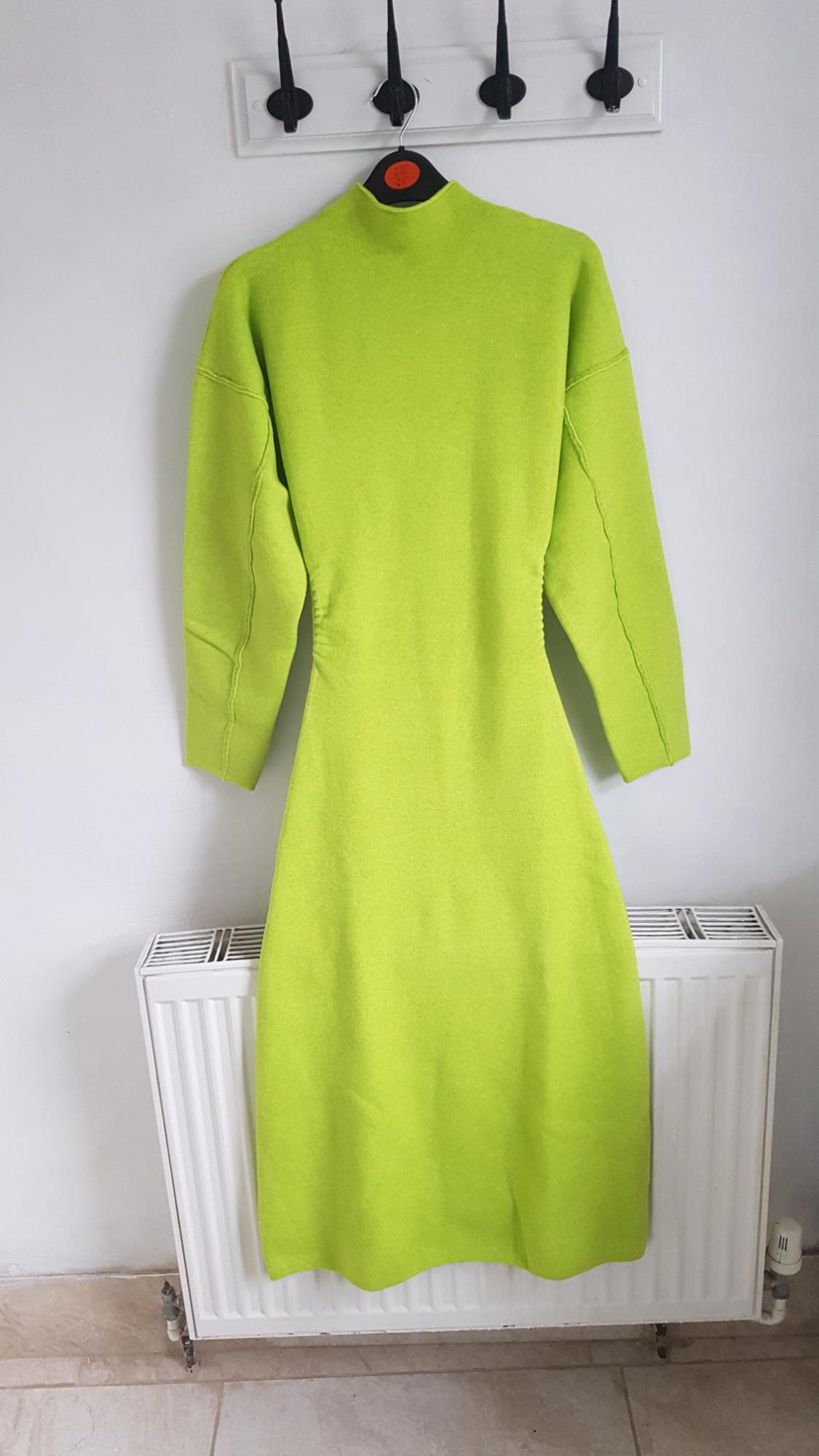 Lime green, long-sleeved dress hanging on a black hanger over a white radiator, on a white wall.