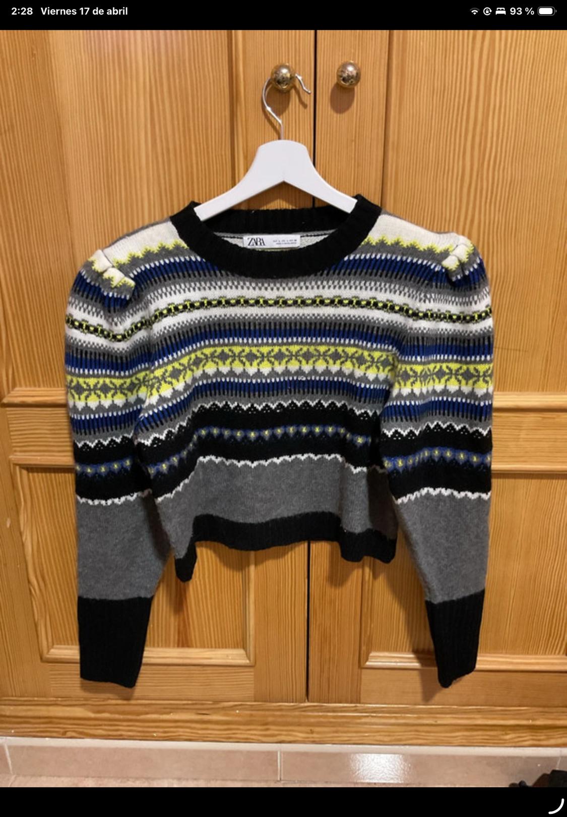 Knitted jumper hanging on a white hanger, with a striped and patterned design in grey, blue, black, and yellow, in front of a wooden door.