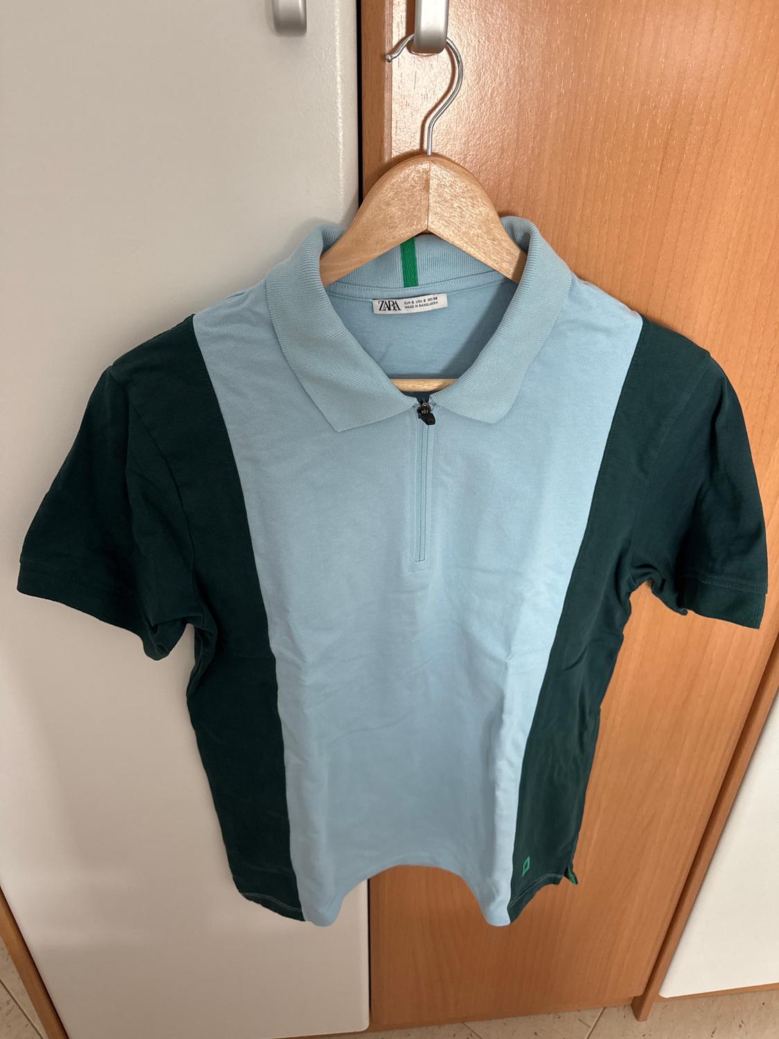 Two-tone shirt hanging on a wooden hanger. The central part is light blue and the sides are dark green. The label is visible on the collar.