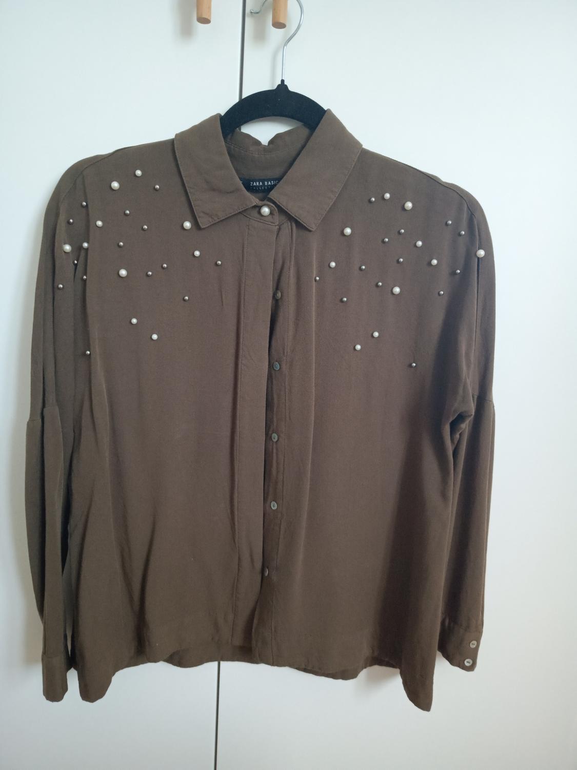 Brown shirt with pearls and studs, hanging on a black hanger in front of a white wardrobe. Text on label: ZARA BASIC.