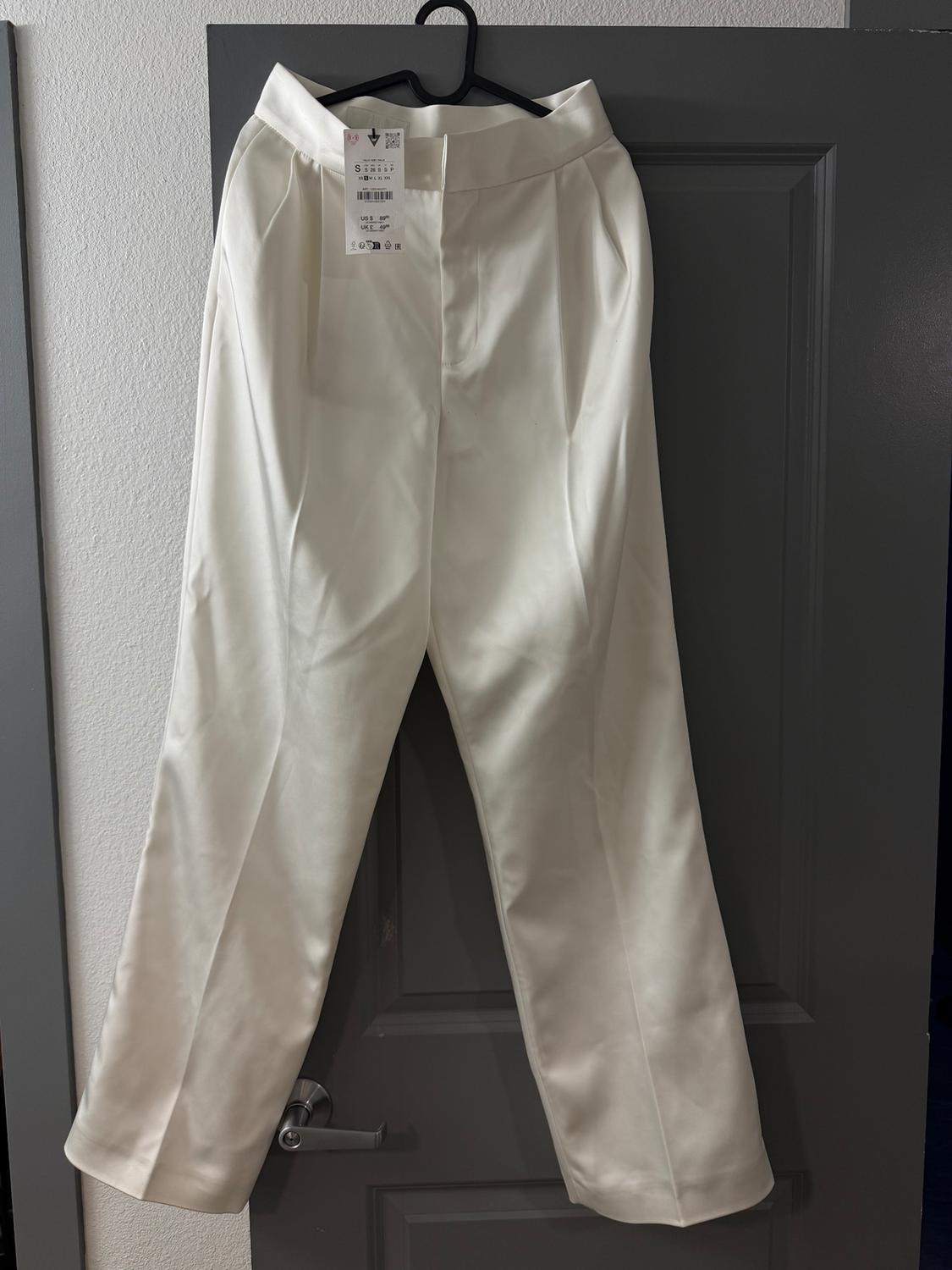 White trousers hanging on a grey door, with a visible tag showing product details. Indoor setting with soft lighting.