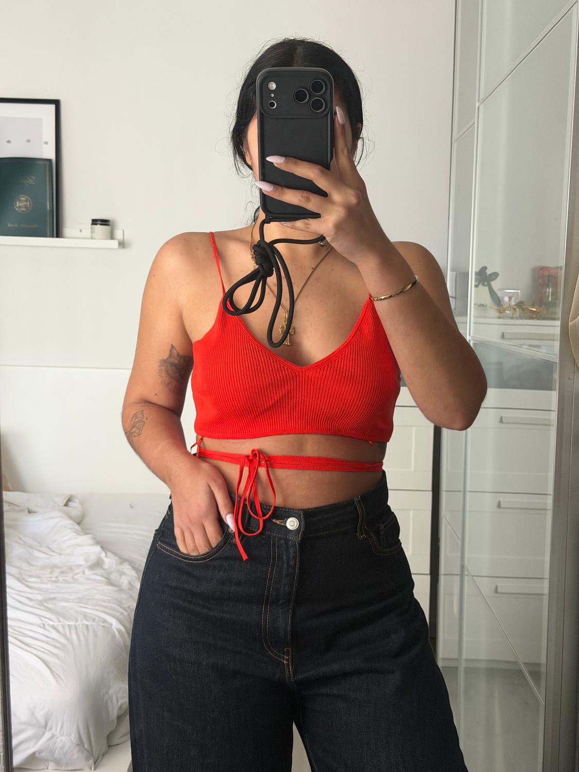 Woman in a red spaghetti-strap top and dark jeans, taking a selfie in a mirror. White room with an unmade bed in the background.