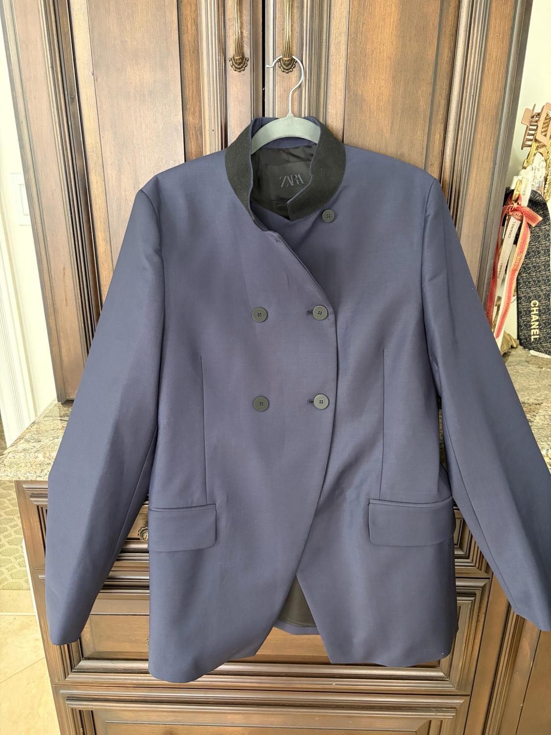 Navy blue double-breasted jacket with a black collar, hanging on a hanger in front of a wooden wardrobe. Text on the label: ZARA.